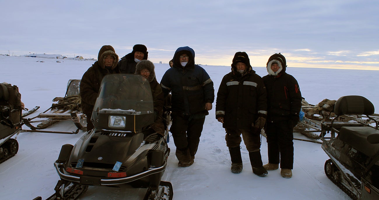 A group of men sit and stand around snow machines somewhere in the Arctic with snow.