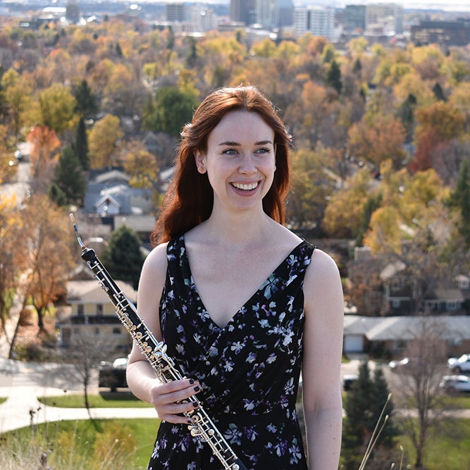 Photo of Rachel standing outside holding an oboe. Behind her are residential home and autumn colored trees