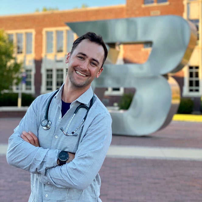Dane Larson smiles in front of the Boise State B wearing his stethoscope around his neck.