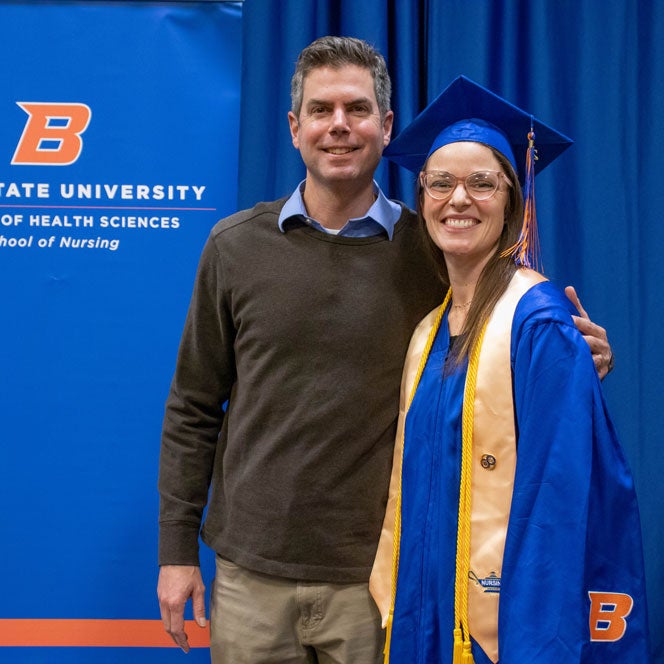 Margaret Quatraro stands in regalia with her husband front of the School of Nursing banner.