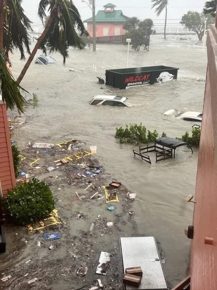 Flood waters cover the streets during a storm, filling them with debris and floating vehicles.