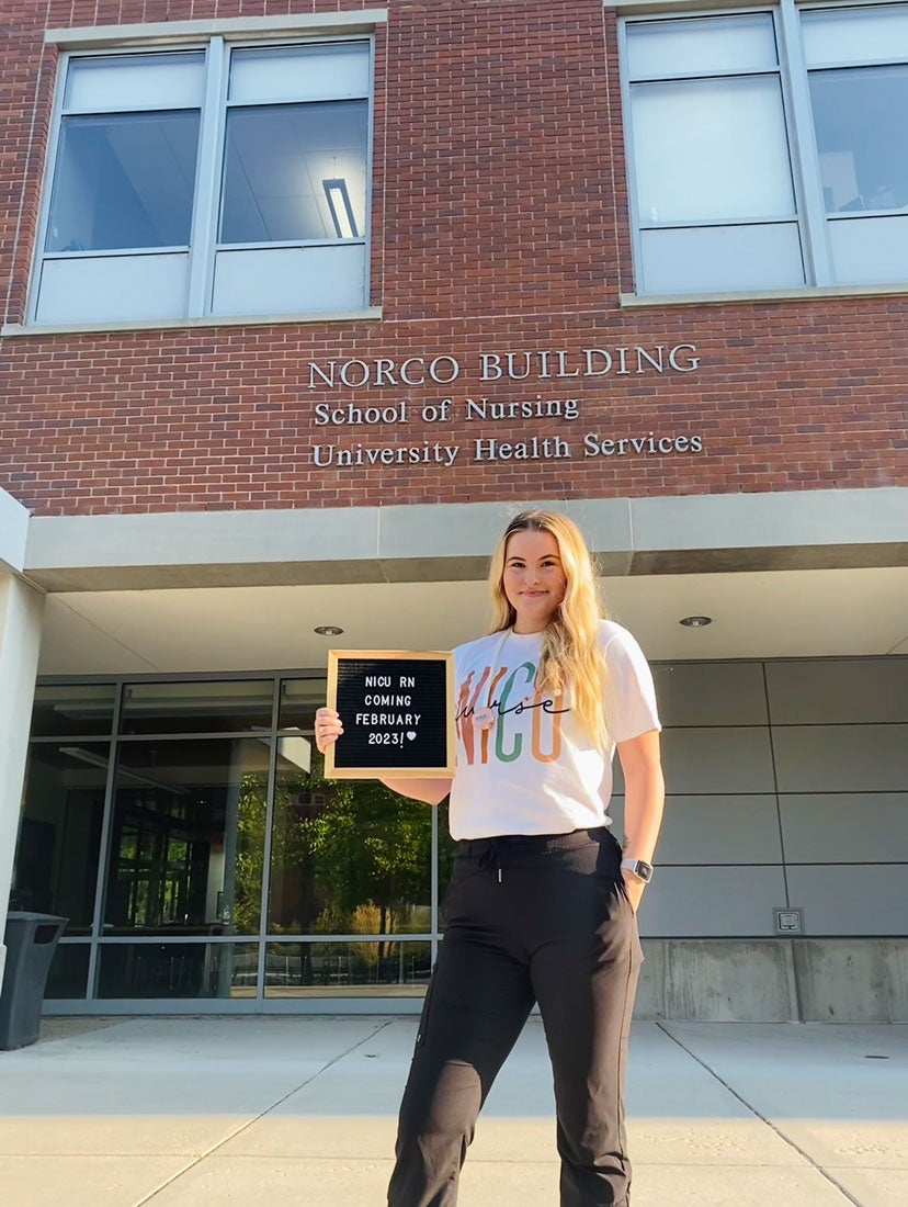 Amanda Pace stands in front of the Norco Building holding a sign that reads: NICU RN coming 2023!