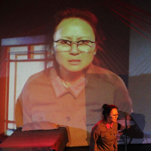 Lo scrutinizes a camera that projects a larger image of herself in the background