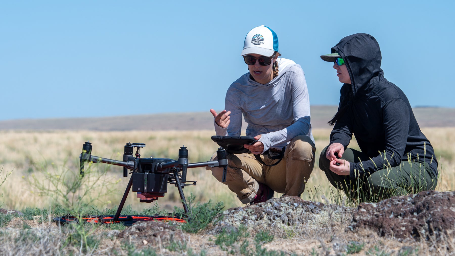 two people crouch on ground beside a drone