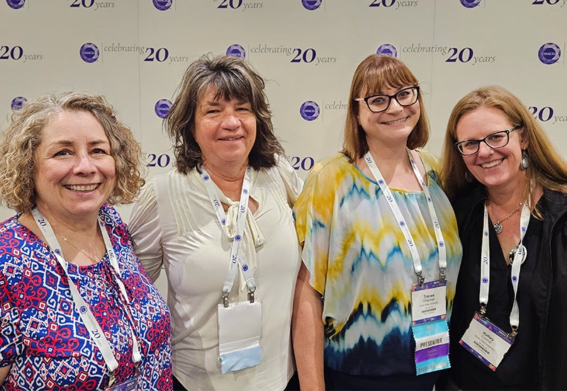 Paula Molina-Shaver, Tammie Sherner, Tracee Chapman and Kelley Connor stand together in from of a conference banner.