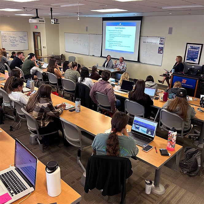 A full lecture hall of students listens to two guest students in the front of the room.