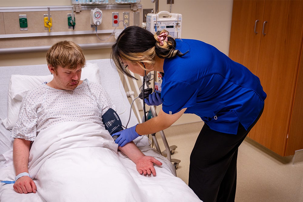 A patient lies in a hospital bed while a nursing student takes their blood sugar.