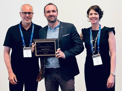 two men and one woman stand for posed photo, man in center hold award plaque