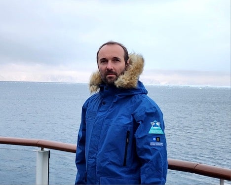 man in puffy winter coat stands on deck of ship, sea and ice behind him