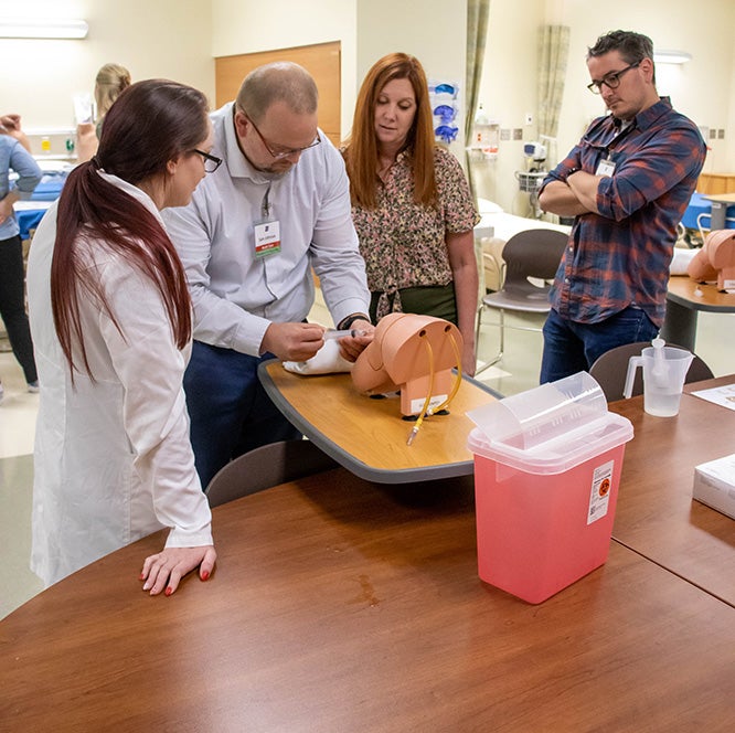 A nurse practitioner student practices giving a joint injection while their professor looks on.