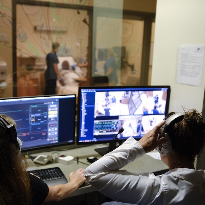 Two educators sit behind computers monitoring a simulation in the other room, slightly visible through one-way glass.