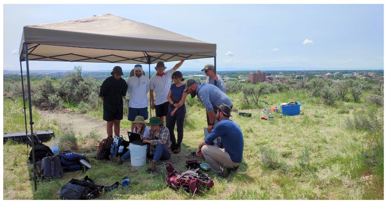 Students work under a canopy, buildings are is visible in the distance