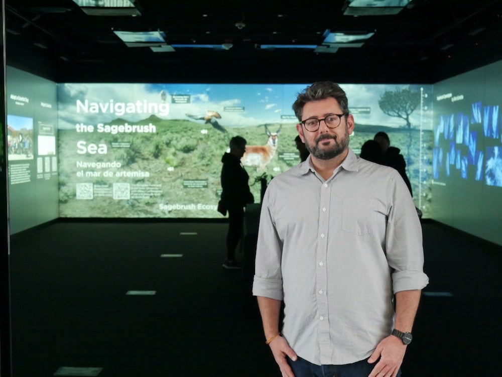 a man poses in front of a projected screen of sagebrush