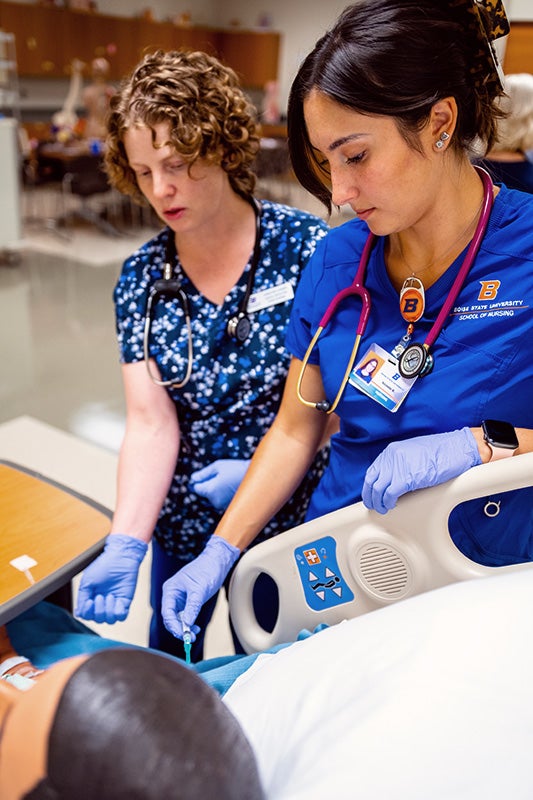 A nurse instructor stands behind a student giving an injection to a manikin.