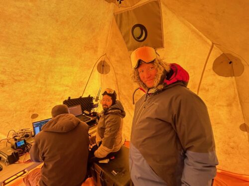 three men in witer coats stand in yellow tent conducting computer work