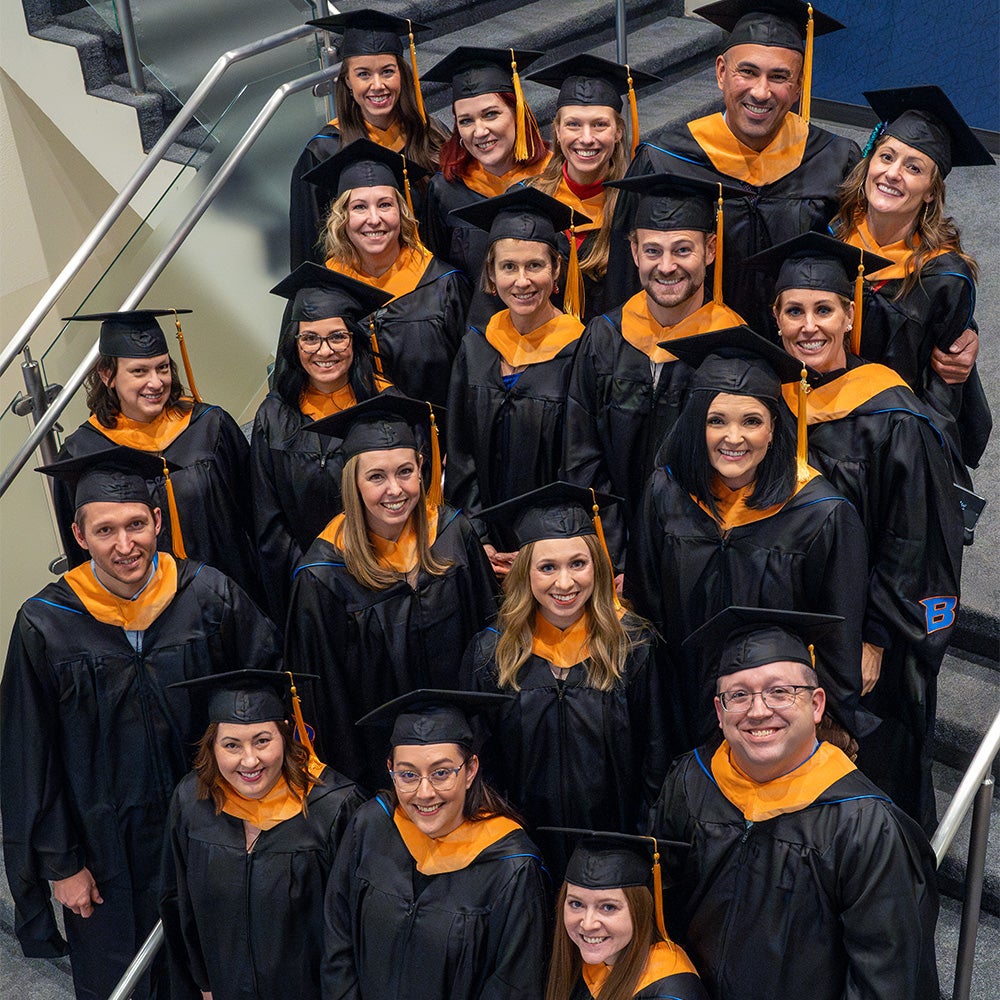 A cohort of AGNP graduates in their regalia stand on a staircase and smile up at the camera.