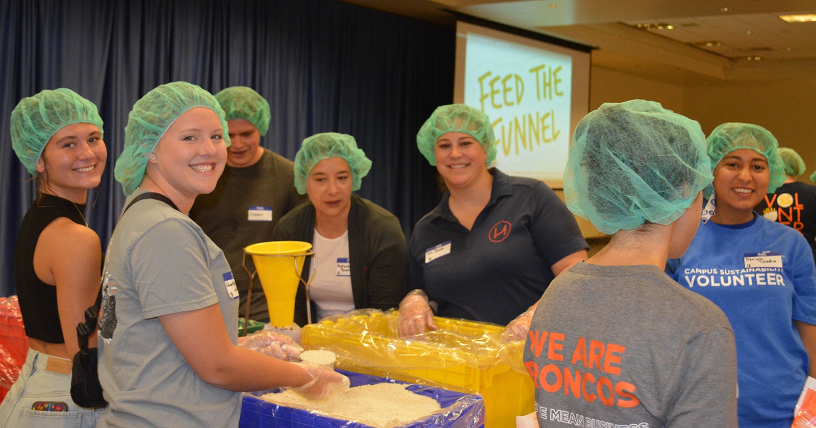 A group of smiling people wearing hairnets and gloves work at a table packing food into containers.