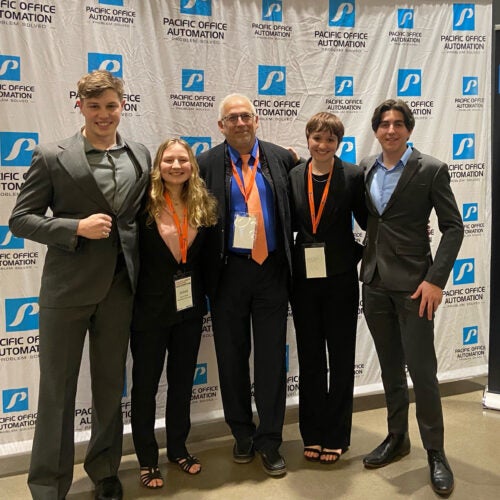 Group of four students and their instructor dressed in business clothes stand in front of a backdrop at a sales competition.