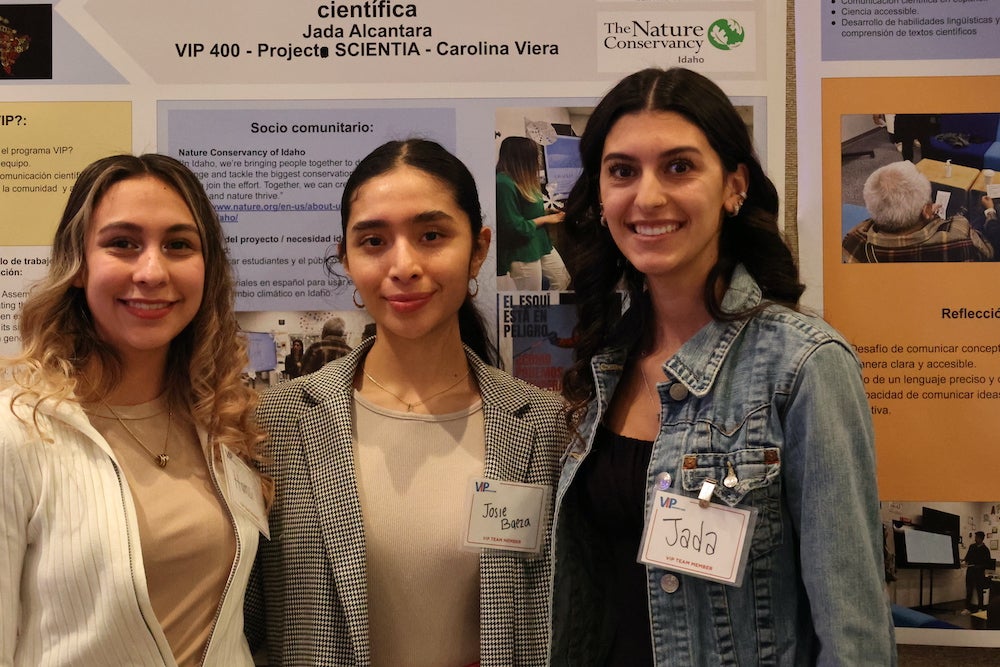 three women link arms in photo. they are standing in front of a research poster