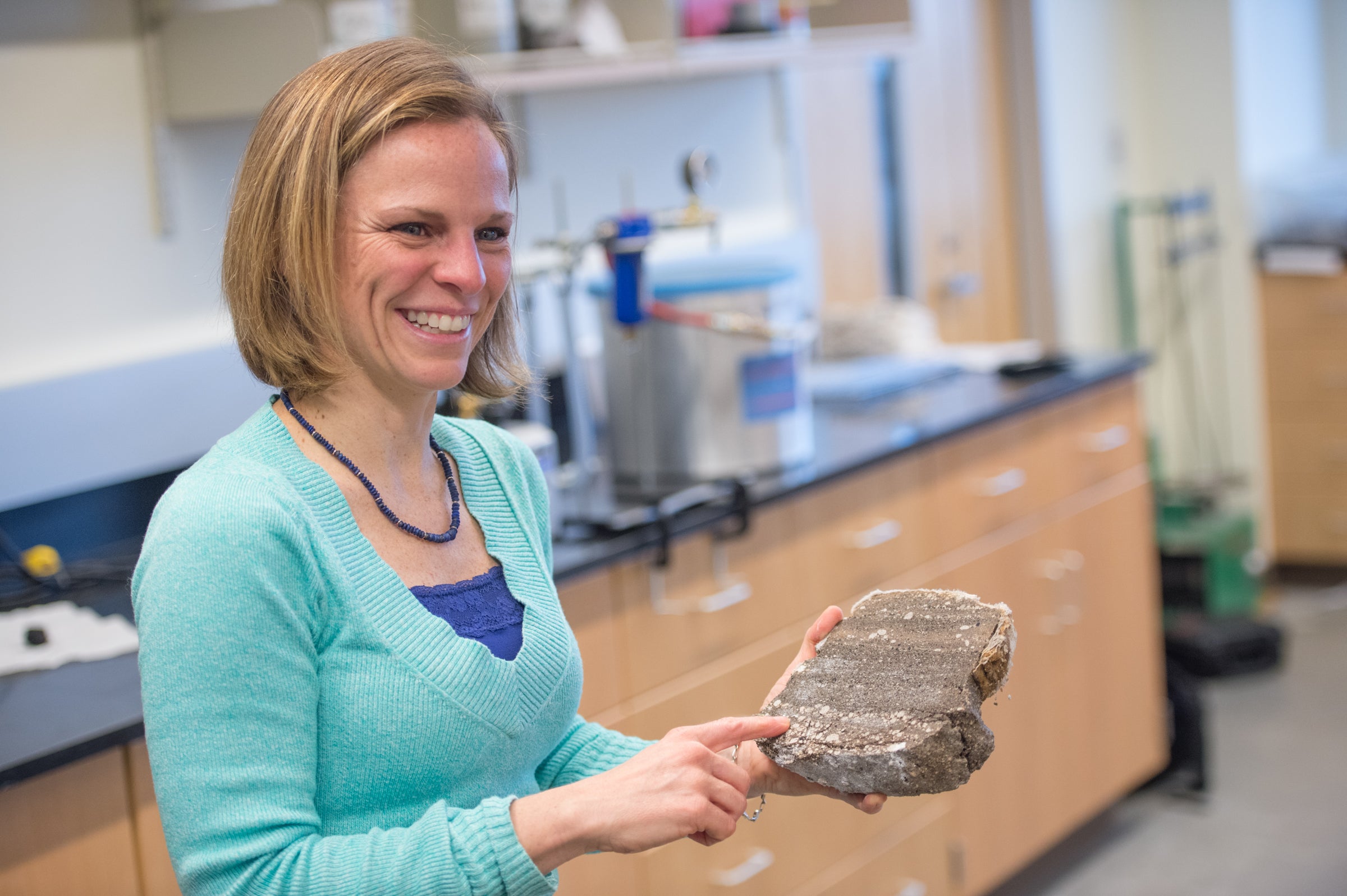 Brand is pictured in a science lab with a volcanic rock. She is smiling in a cyan sweater and is pointing to the rock.