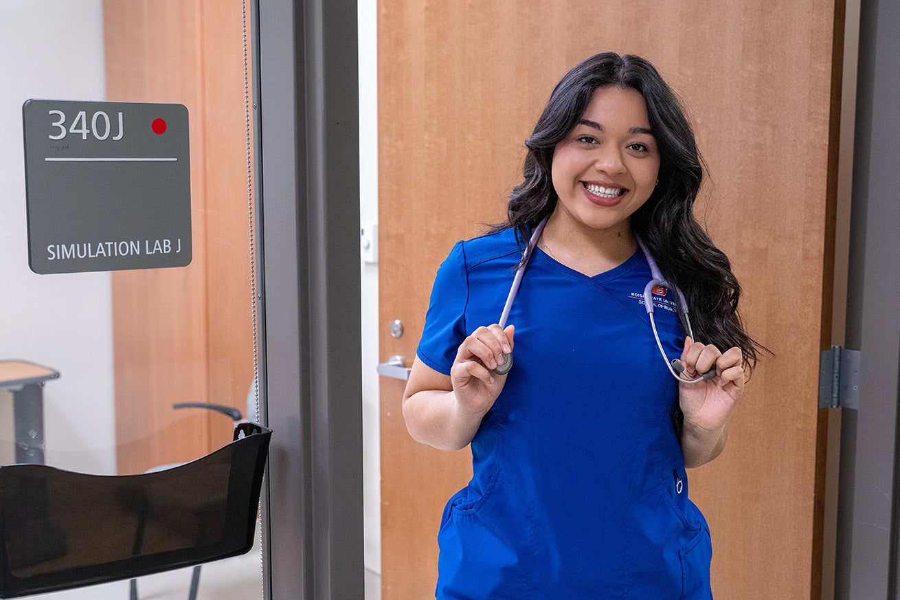 A nursing student wears blue scrubs and a stethoscope around her neck. She stands in the door of a simulation patient room and smiles.