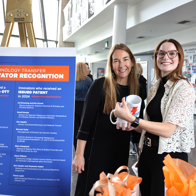 Nancy Glenn and Sarah Anderson stand near a poster and hold OTT personalized event mugs.