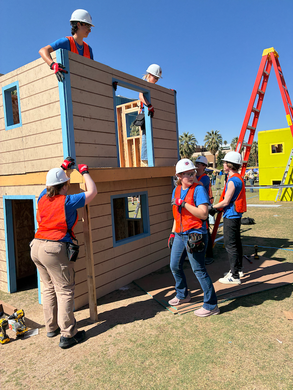 six students in orange high visibility vests work on a miniature two story timber structure