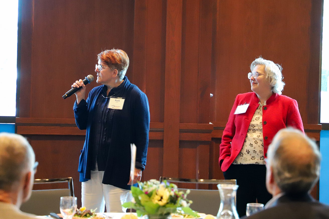 A woman in navy speaks into a microphone while a woman in a red blazer smiles.