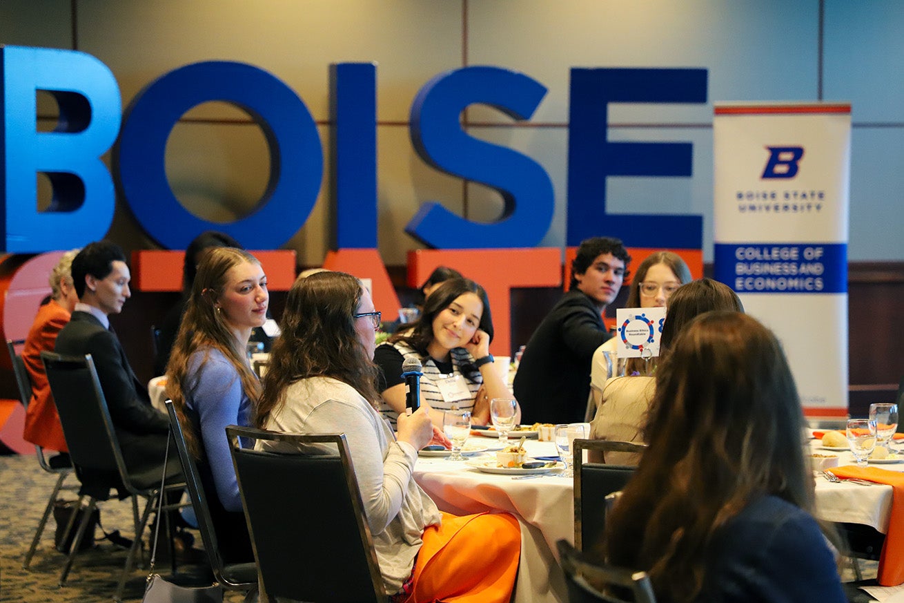 Students sit around tables set for dinner and listen to one student in the foreground who holds a microphone.