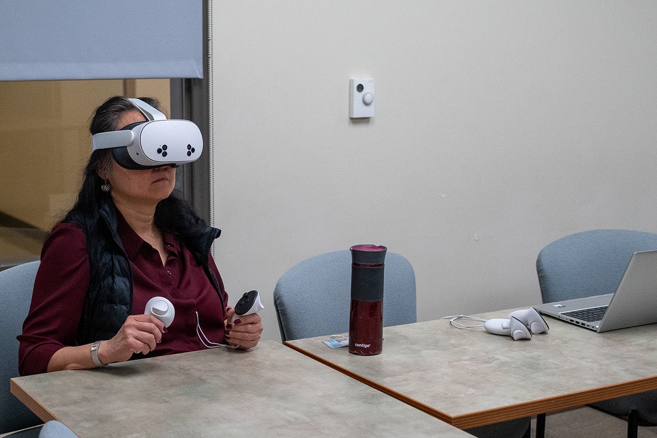 A woman sits at a table while holding virtual reality hand controllers and wearing a headset over her eyes.