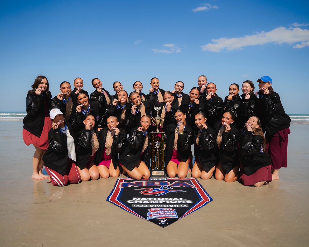 Boise State Mane Line dancers biting their medals and posing with trophy on the beach.