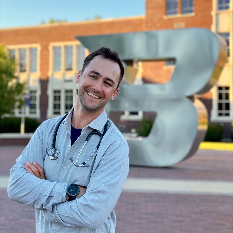 Dane Larson stands in front of the Boise State B smiling with his stethoscope around his neck.