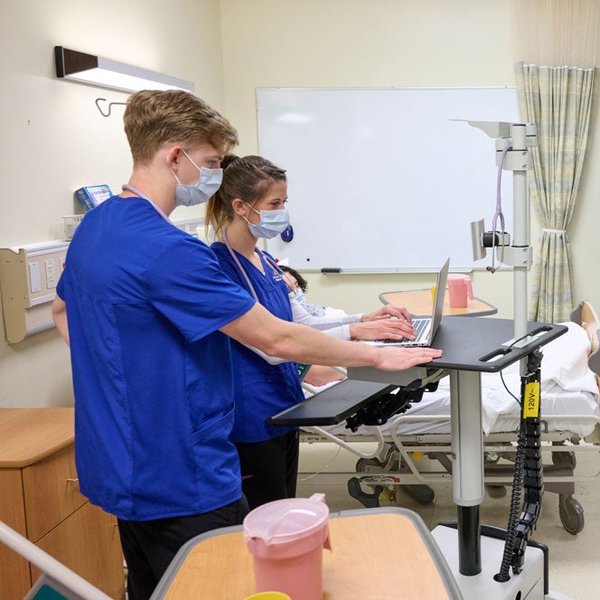 Nursing students in blue scrubs input data into a laptop in the skills lab.