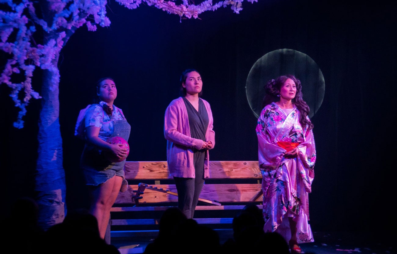 Janet Lo stands in blue lighting on a stage with two other women.