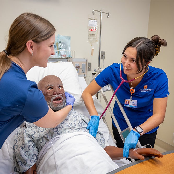 A student uses a stethoscope to listen to a manikin's lungs as another student adjusts his oxygen tube.