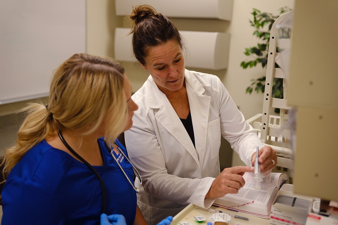 A professor in a white coat points to the label on a syringe as she talks to a student in scrubs.