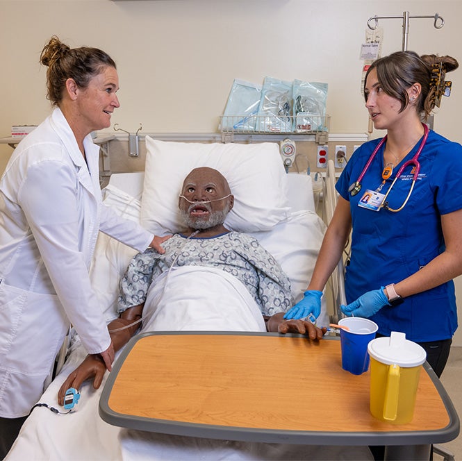 A professor in a white coat stands on one side of a manikin in a bed and a student in scrubs stands on the other.
