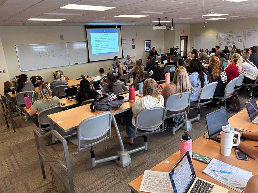A full lecture hall of students listens to two guest speakers at the front of the room.