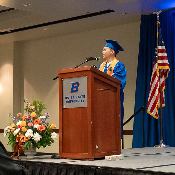 Jan Bobadilla stands on stage at the podium in his blue and orange regalia.