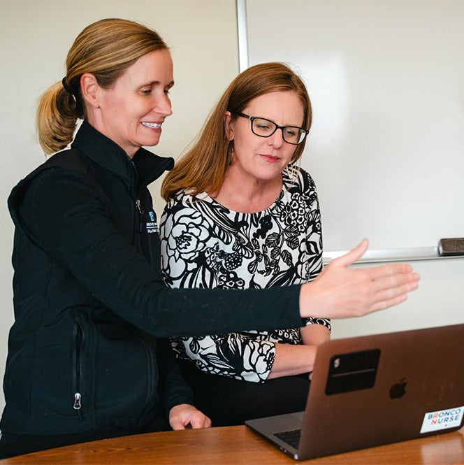 Kelley reads Jenny's laptop screen as Jenny motions with her hand to explain what she's looking at.