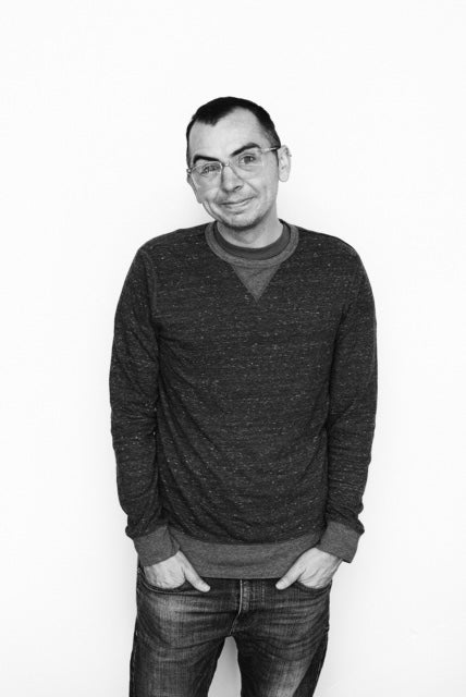 Black and white photo of Tate Klacsmann standing with his hands in his pockets smiling.