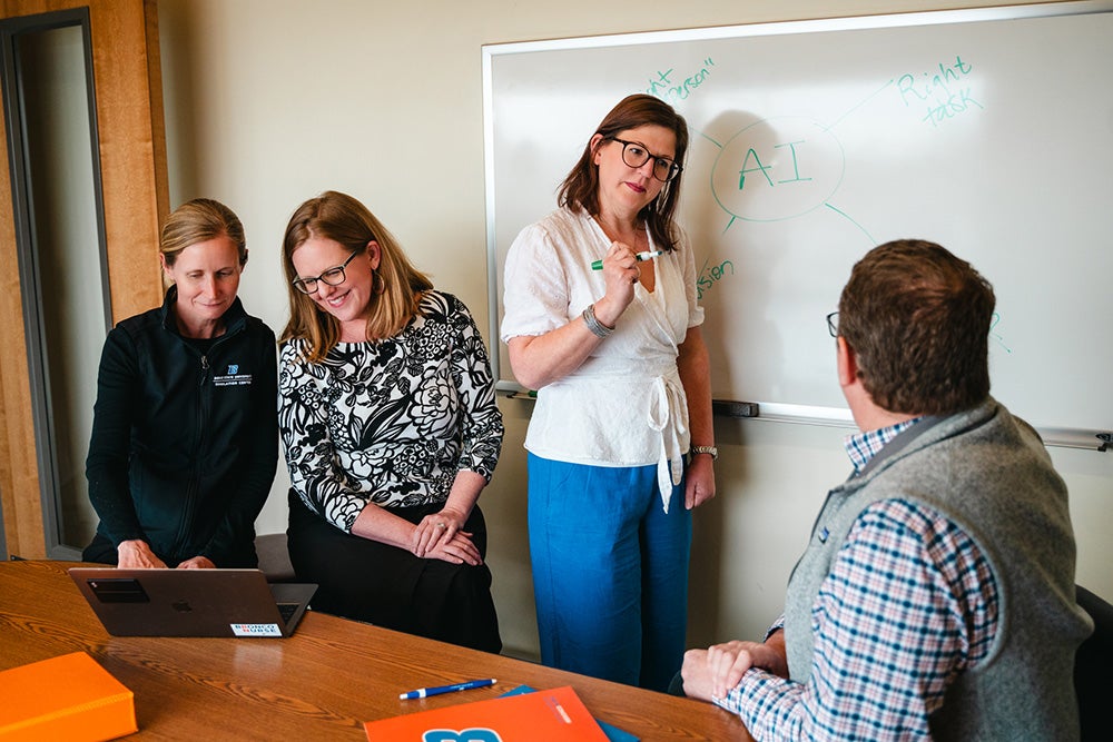Jenny and Kelley look on to a laptop while Sarah stands at a white board and talks with Jason who is sitting at the table.