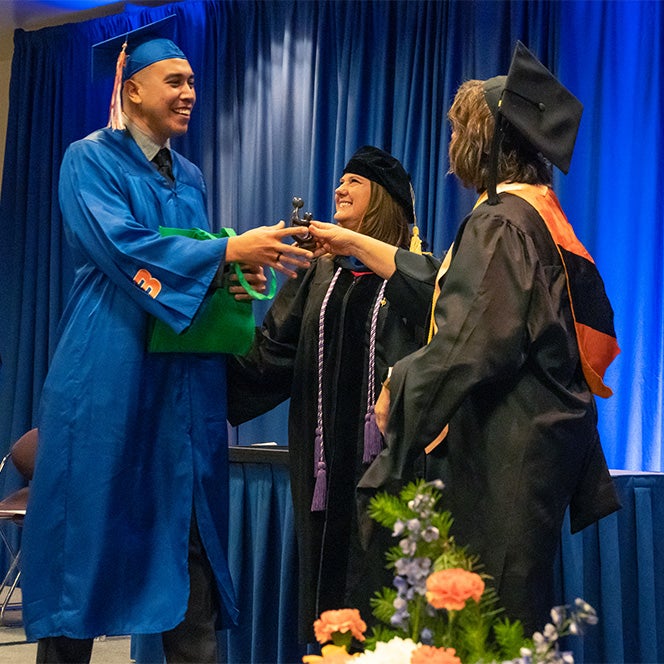 A student in regalia accepts a small, dark grey sculpture from two faculty.