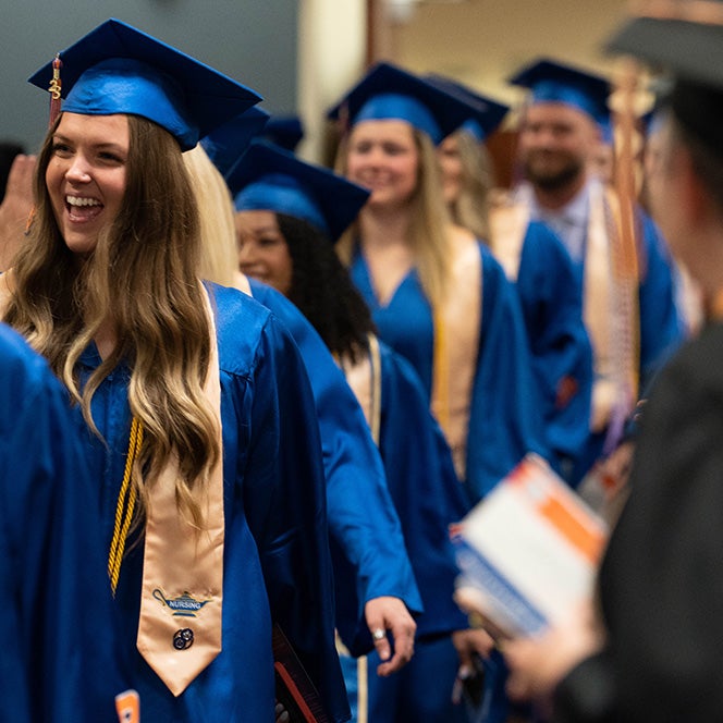 Nursing students walk in a row during Convocation in their caps and gowns. In the foreground is the a stole with the lamp design and nursing pin.