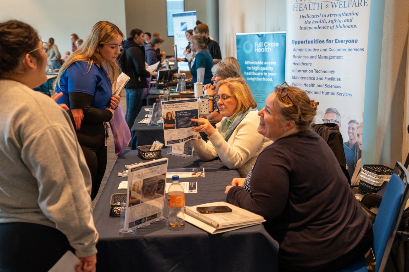 Students talk to two smiling people behind a table and look at posters and fliers advertising healthcare careers.