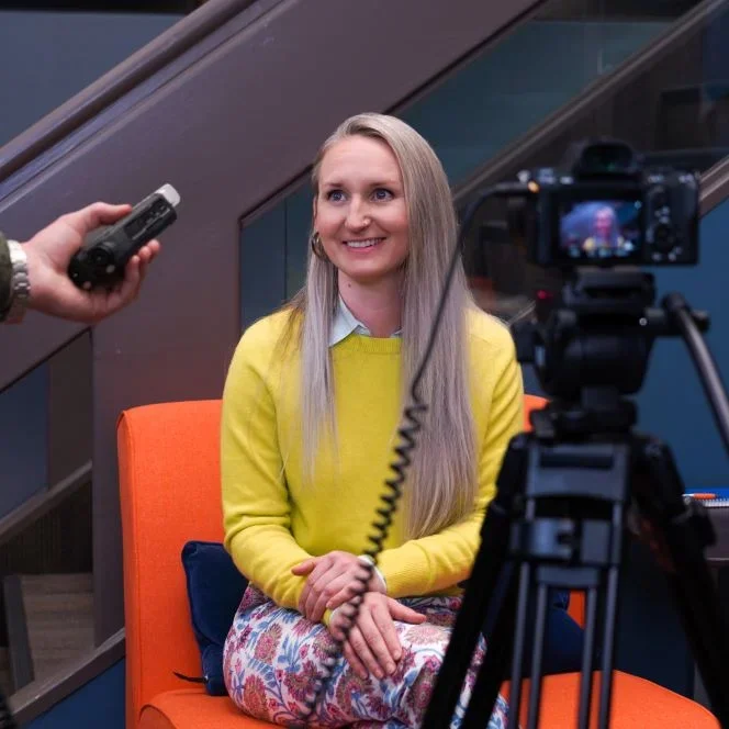 Jessi Boyer sits on orange chair and in front of a camera.