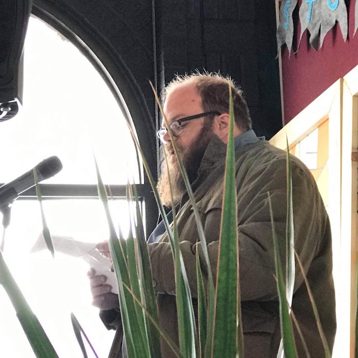John Barrie at a reading. A bright window is in the background and plants in the foreground.