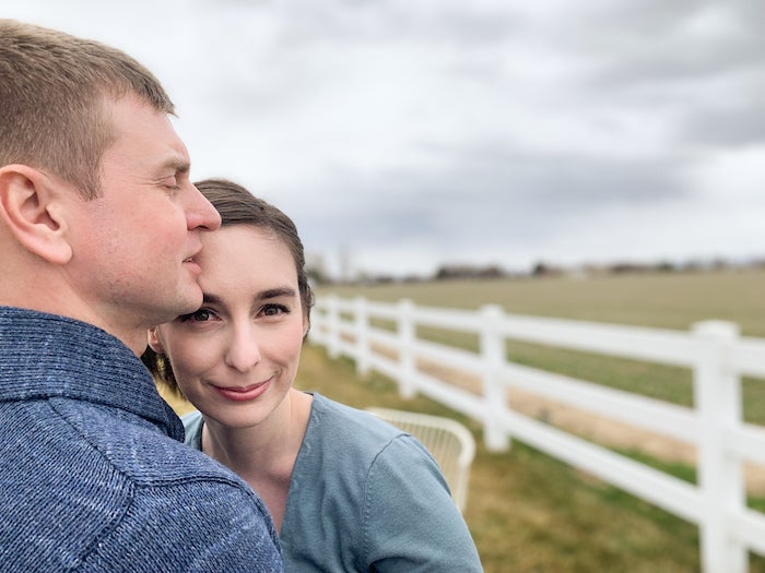 Pania leans against her husband. A cloudy sky, a white fence, and fields are in the distance.