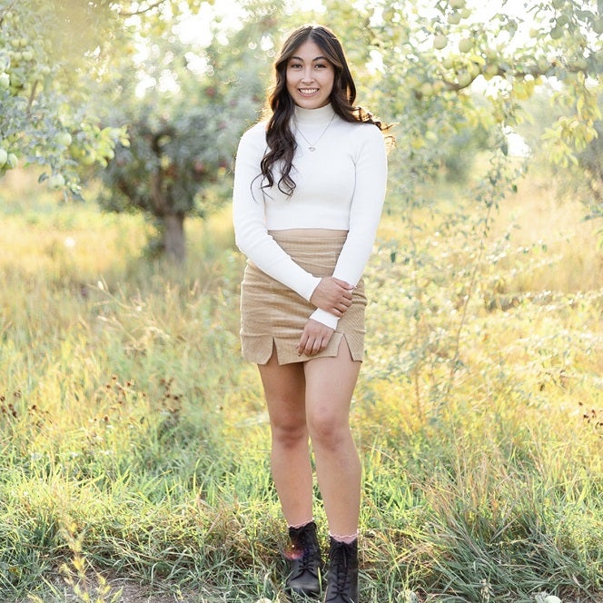 Maiah stands in the middle of an apple orchard, sun shines through the trees