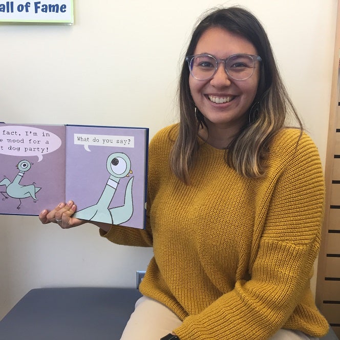 Nicole, a brown haired girl wearing glasses holding open a copy of Mo Willem's Don't Let the Pigeon Drive the Bus